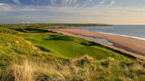 A coastal golf course with a beach view, grassy areas, and a flag marking the hole. - Wilde Ballybunion (Ballybunion)