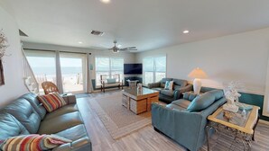 A spacious living room with a large sofa, a coffee table, and a television. There is a balcony with a view of the ocean. - Spacious 4 Bedroom Gulf Front Home with large deck overlooking Gulf of Mexico (Port Saint Joe)