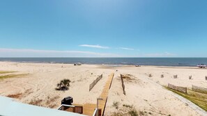 Vista a la playa desde un balcón con terraza de madera, playa de arena y cielo despejado.