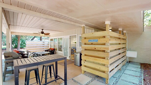 Ein überdachter Außenbereich mit Holztisch und -stühlen, einer Holzpallete mit der Aufschrift 'Dusche' und einem Kühlschrank.