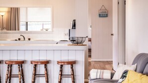 A kitchen with a white countertop, three wooden stools, a hanging sign that says "LOVE THE BEACH", and a door leading to another room. - Stones Throw - Holiday Houses Tasmania (Primrose Sands)