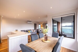 Een moderne keuken met een wit aanrecht, grijze bekleden zitplaatsen en een eethoek met een houten tafel en grijze stoelen. Een groot raam biedt uitzicht op de zee.