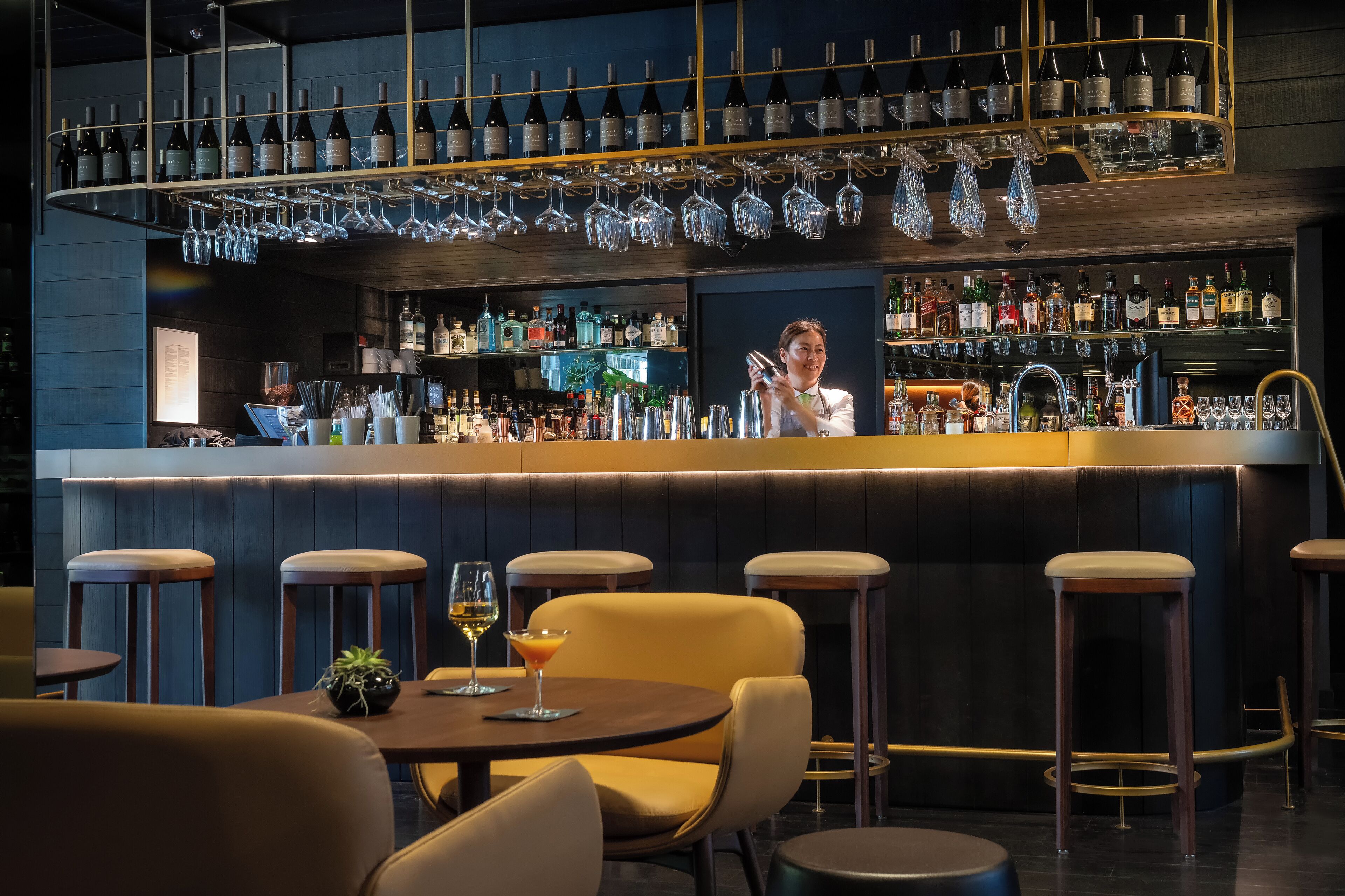 Zona de bar con varios taburetes, una barra con un bartender y una variedad de botellas y vasos.