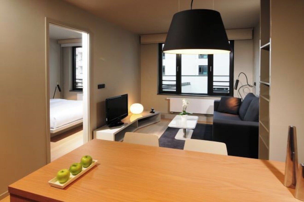 A modern hotel room with a bed, a TV, a dining table with apples, a sofa, and a large window.