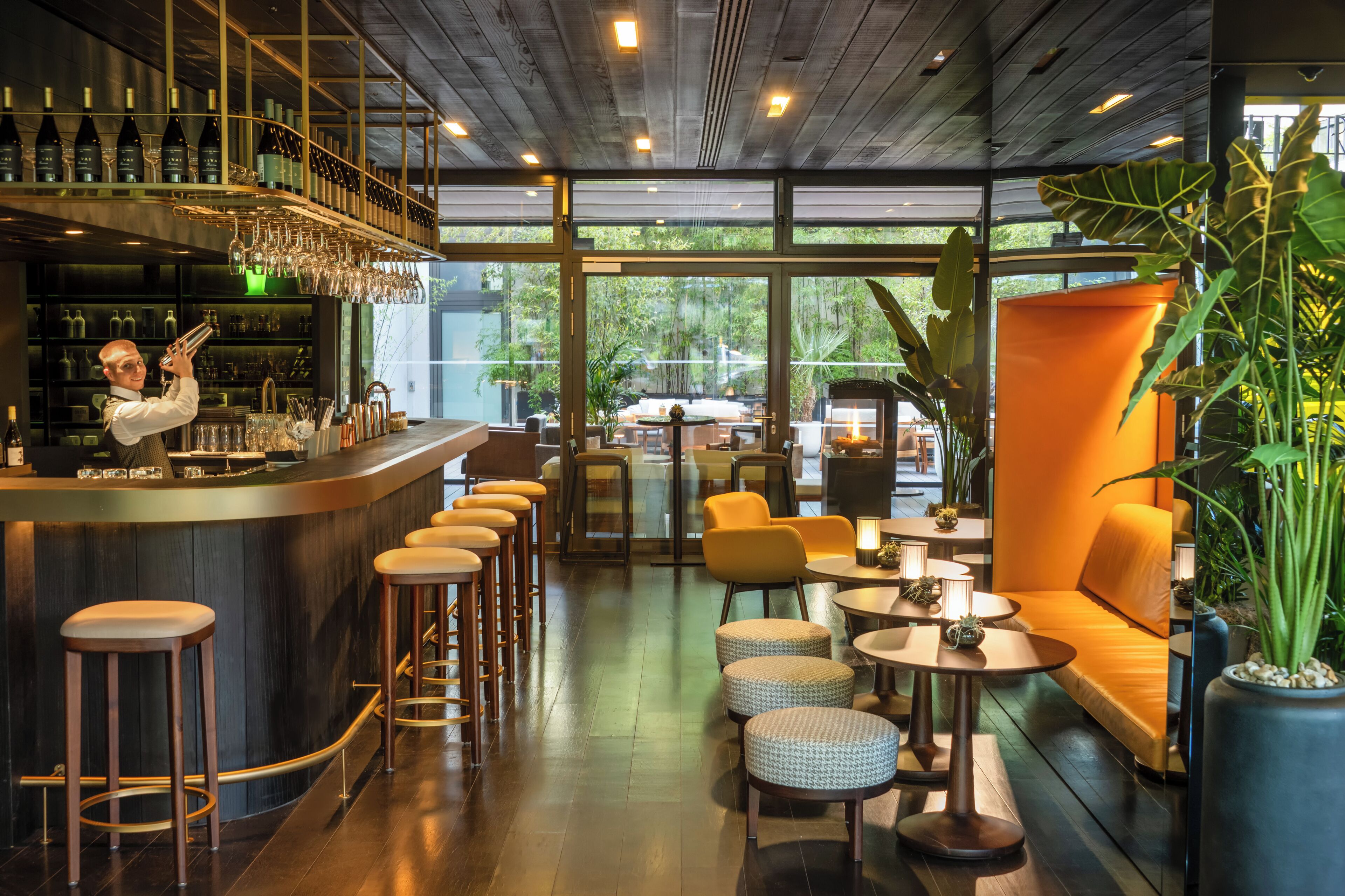 A modern bar area with a bartender, bar stools, and a wine rack. There are seating areas with tables and chairs, and large windows offering a view of the outdoors.