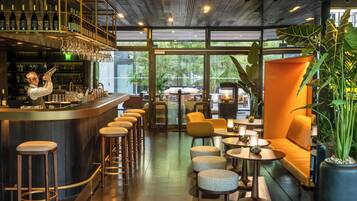 A modern bar area with a bartender, bar stools, and a wine rack. There are seating areas with tables and chairs, and large windows offering a view of the outdoors.