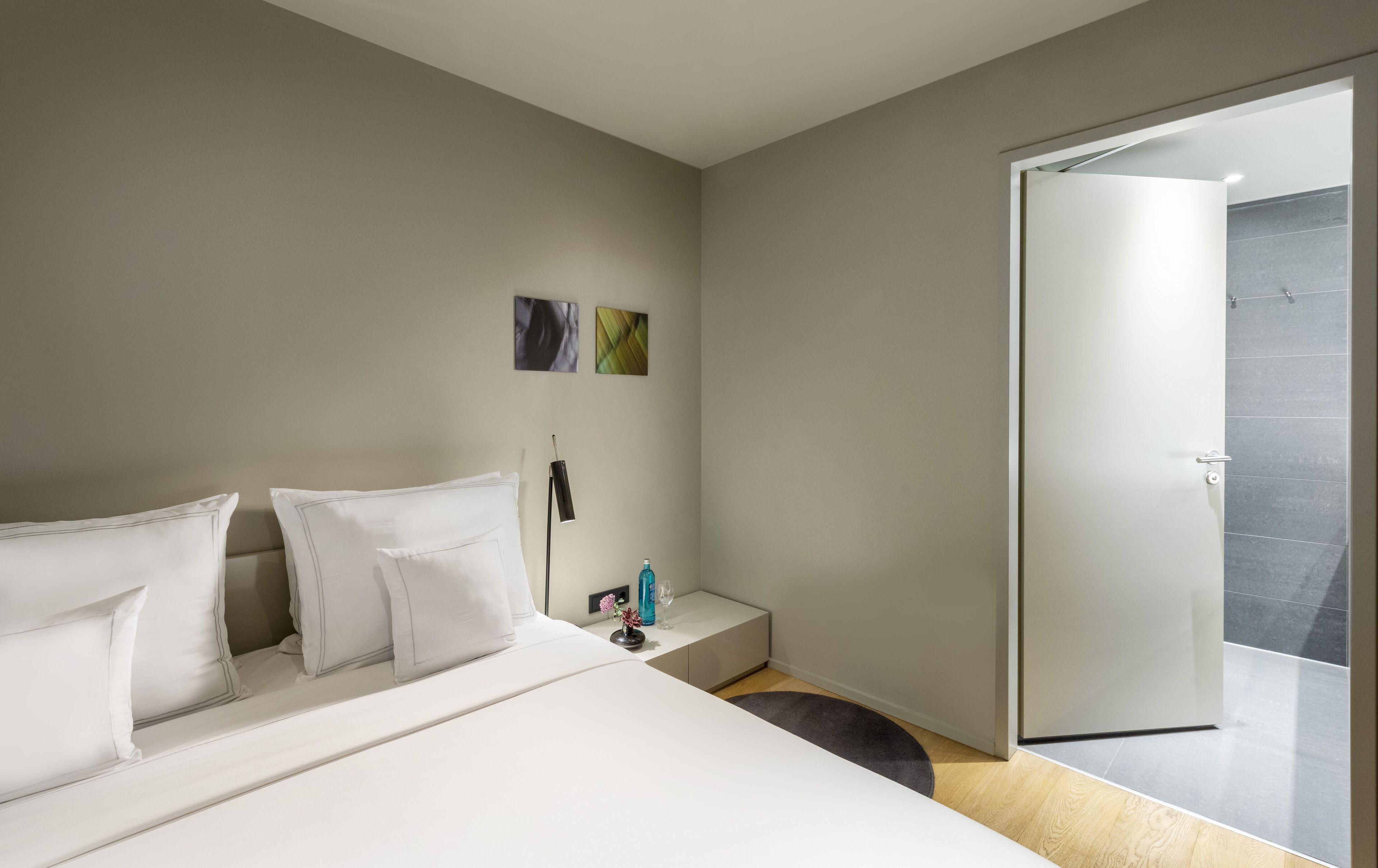 A modern hotel room with a large bed, a bedside table with a lamp, and a view of the outside through a glass door.