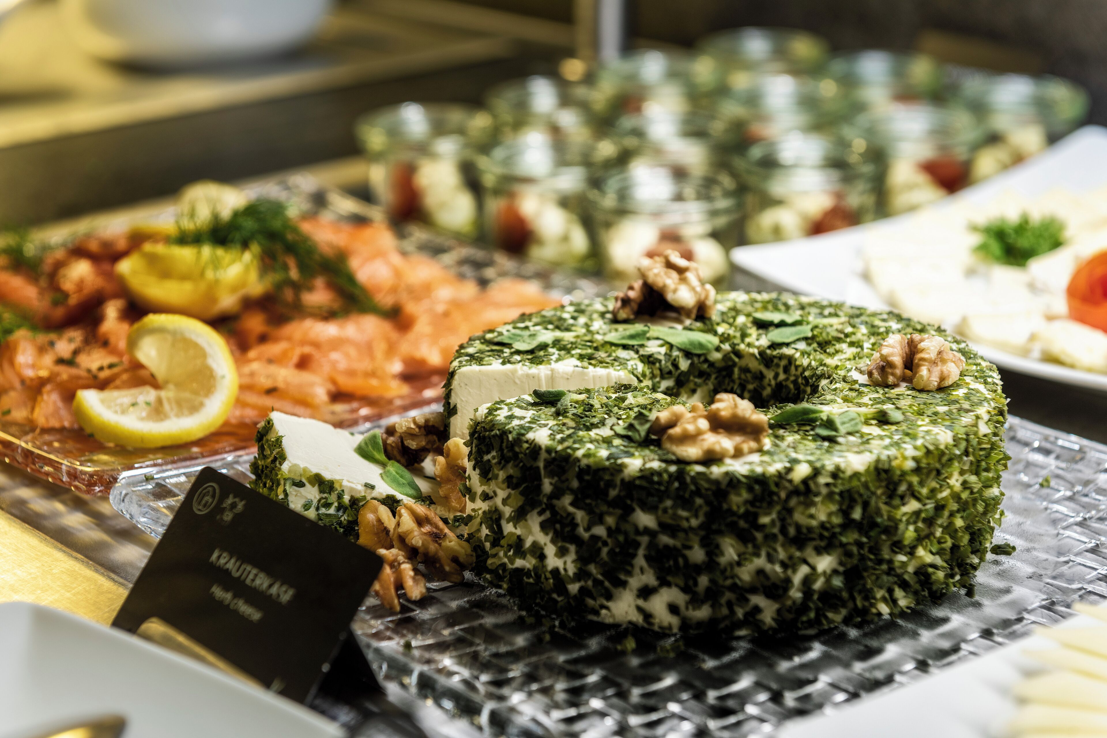 A buffet with various dishes including a green cheese wheel, salmon, and nuts.