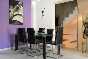 A dining area with a glass table and black chairs, a wall clock, and a staircase with a poster.