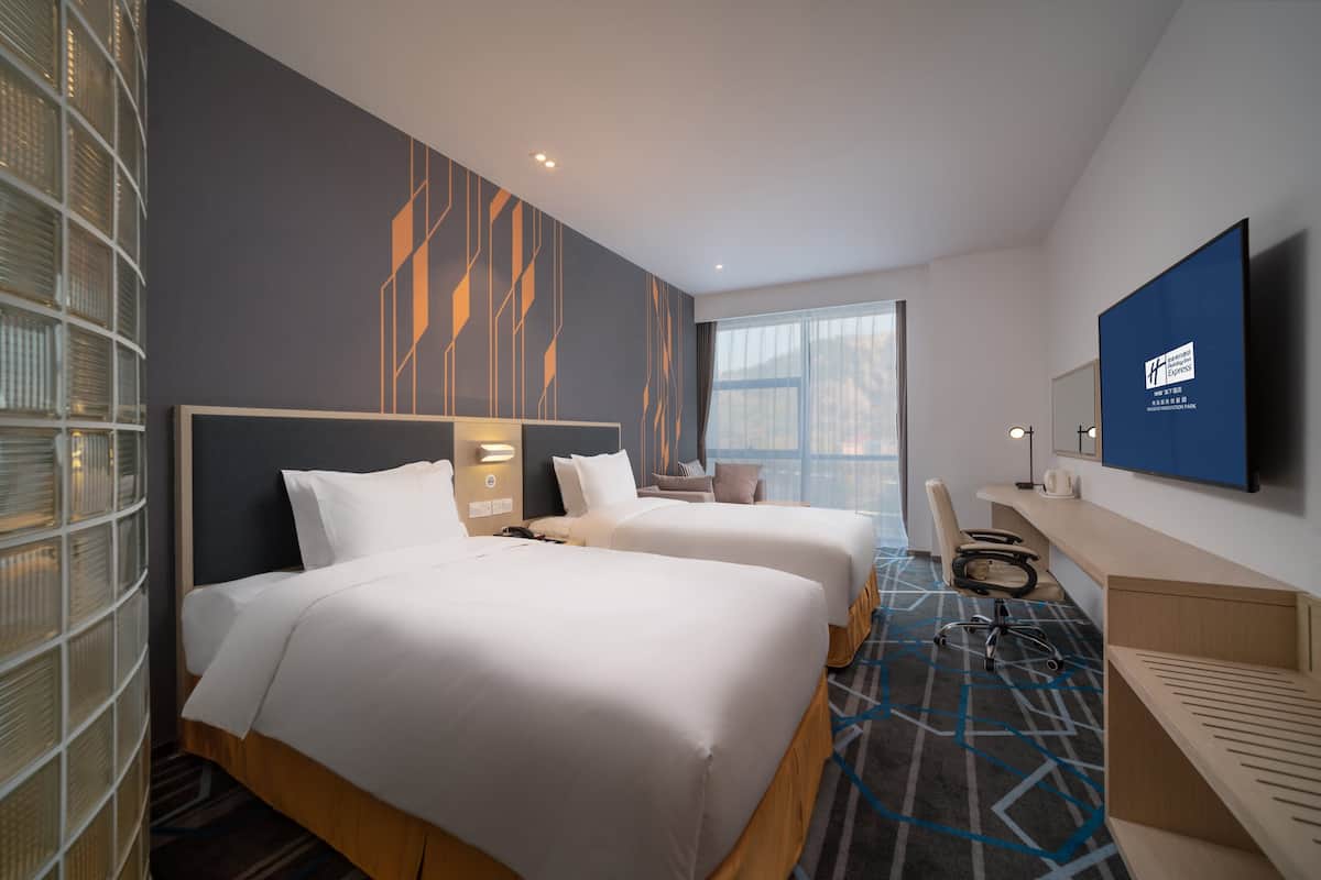 A hotel room with two beds, a desk, a chair, a TV, and a large window.