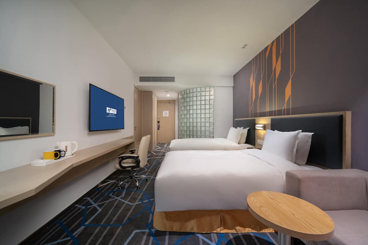 A hotel room with a bed, a desk, a chair, a small table, and a wall-mounted TV.