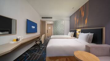 A hotel room with a bed, a desk, a chair, a small table, and a wall-mounted TV.
