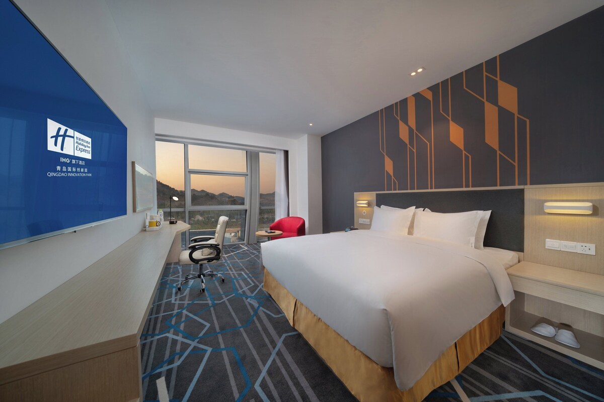 A hotel room with a large bed, a desk, a chair, a red chair, a window with a view, and a wall with geometric patterns.