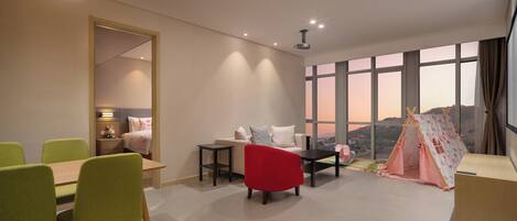 A modern hotel room with a dining area, a bed, a red chair, a small table, a tent, and a large window with a view.