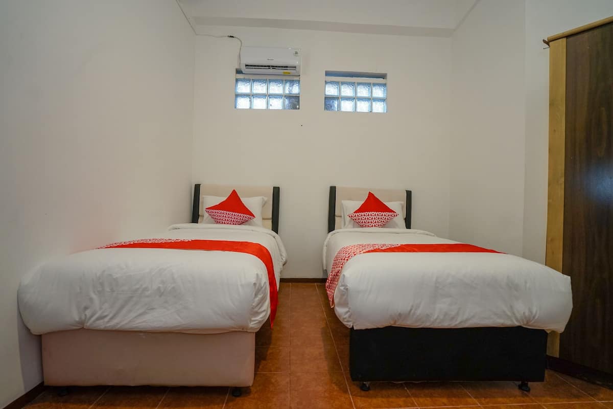 A room with two beds, each with a red pillow and a white bedspread. There is a window with a view of the sky, and an air conditioning unit mounted on the wall.