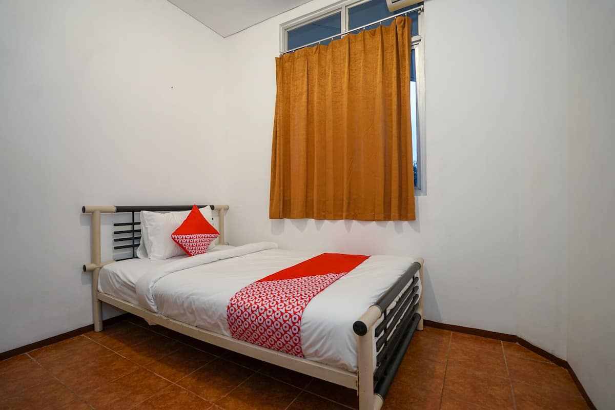 A single bed with a white frame, a red and white patterned bedspread, and a red pillow. There is a window with brown curtains, and the room has white walls and brown tiled flooring.