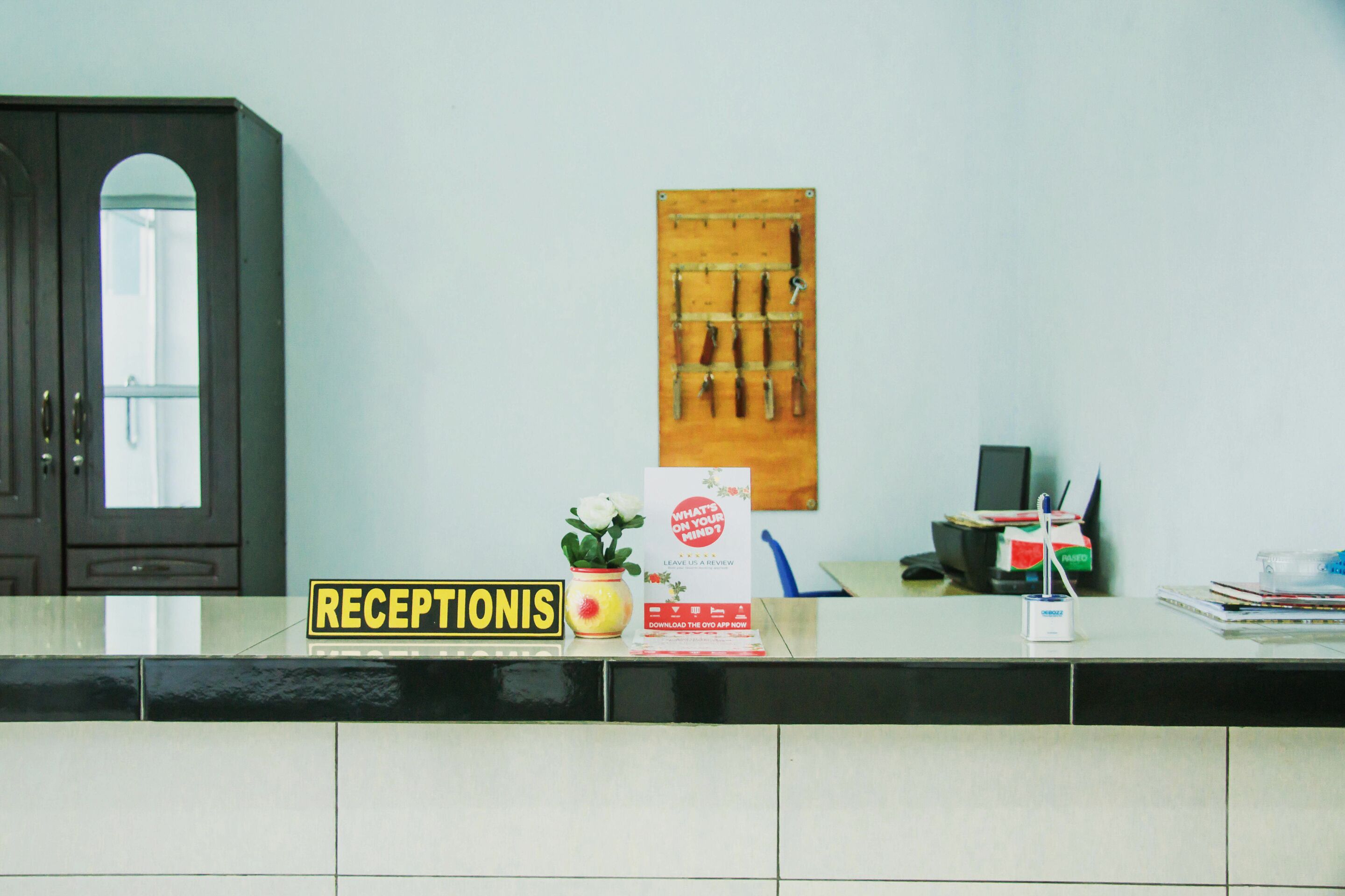 Un área de recepción con un cartel que dice "RECEPTIONIS", una planta, un escritorio con una computadora y un soporte de cuchillos en la pared.
