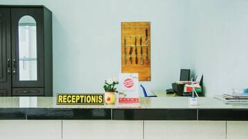 Un área de recepción con un cartel que dice "RECEPTIONIS", una planta, un escritorio con una computadora y un soporte de cuchillos en la pared.