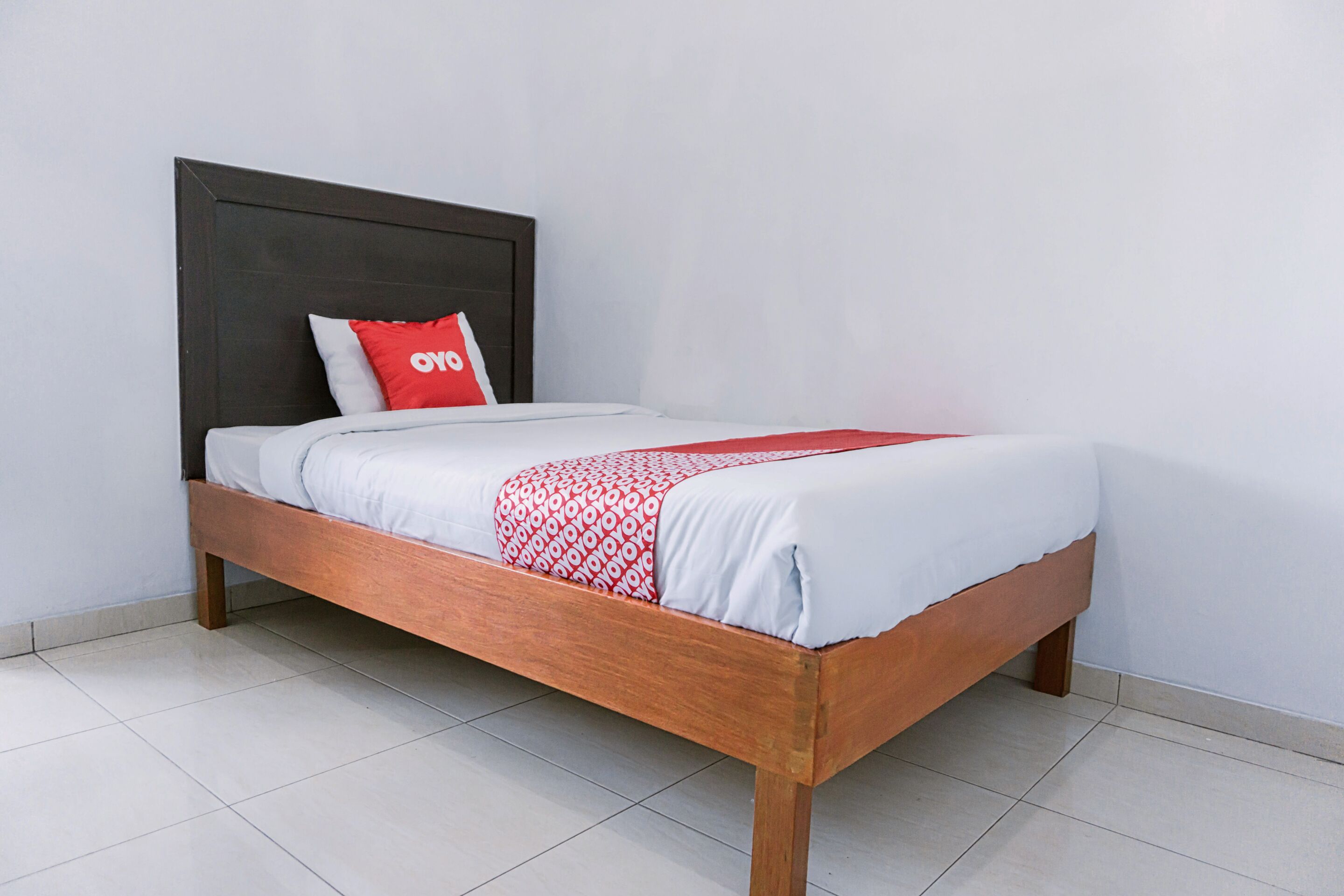 Una cama individual con estructura de madera, ropa de cama blanca y un almohadón rojo con la palabra 'oyo' escrita.