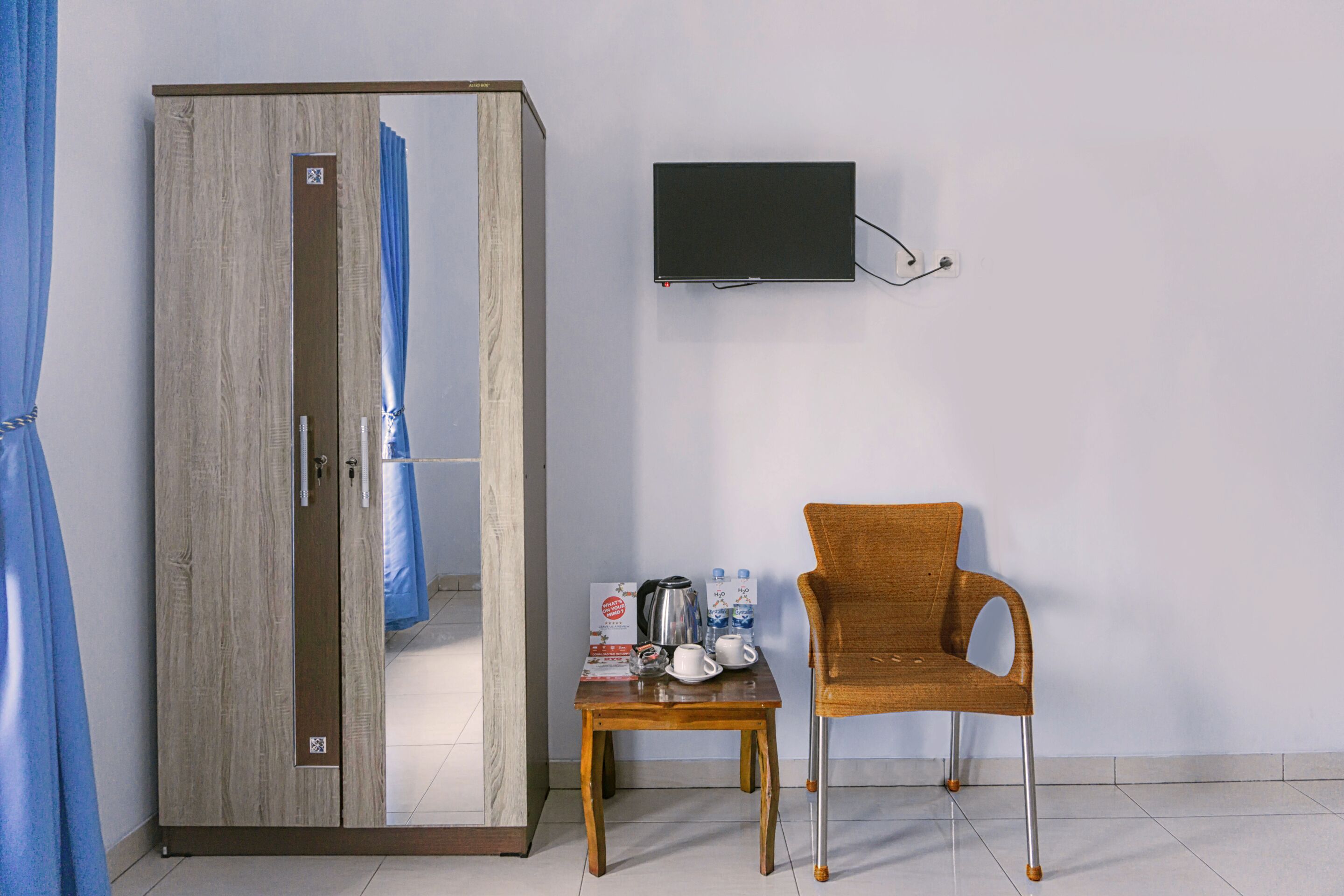 Una habitación con armario, una mesa con silla, televisor de pantalla plana montado en la pared y cortina azul.