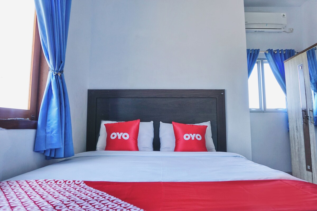 A bed with a white and red bedspread, two red pillows with the word "OYO" on them, and blue curtains on the windows.