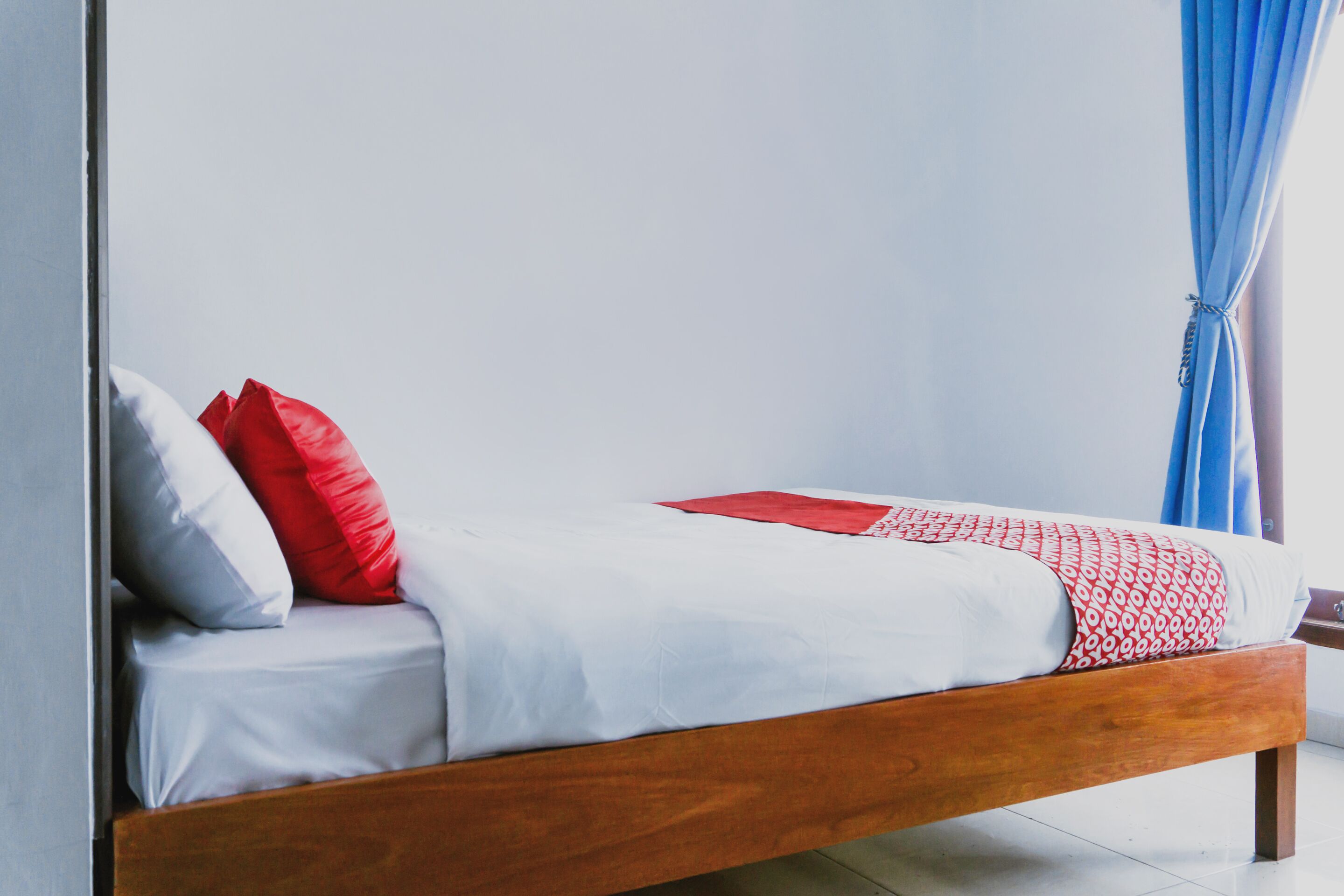A single bed with white bedding and red accents, a wooden frame, and a blue curtain in the background.