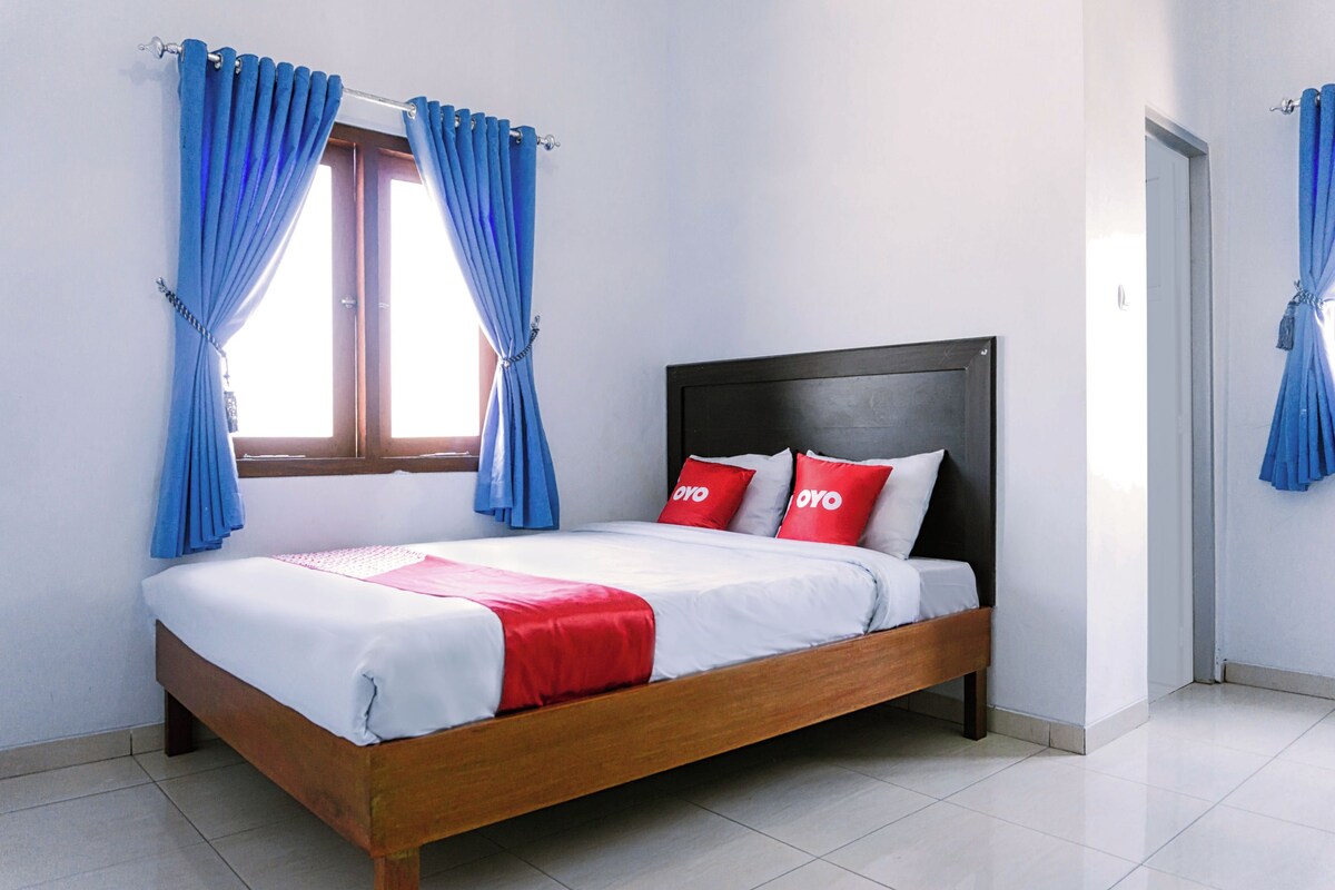 Una cama individual con estructura de madera, ropa de cama blanca y almohadas rojas y blancas. Cortinas azules atadas en la ventana.