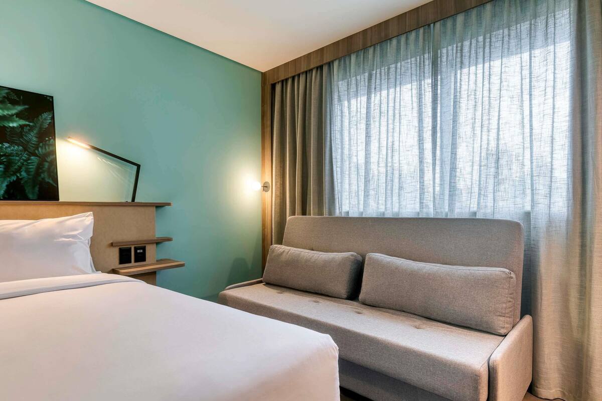 Una habitación de hotel con cama, sofá, estante en la pared y ventana con cortinas.