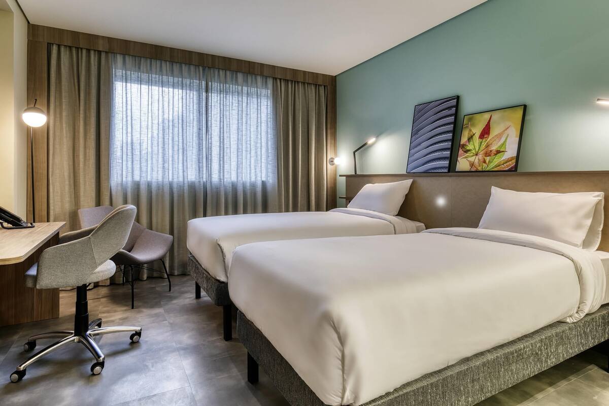Una habitación de hotel con dos camas, un escritorio con una silla, una lámpara y una ventana con cortinas.