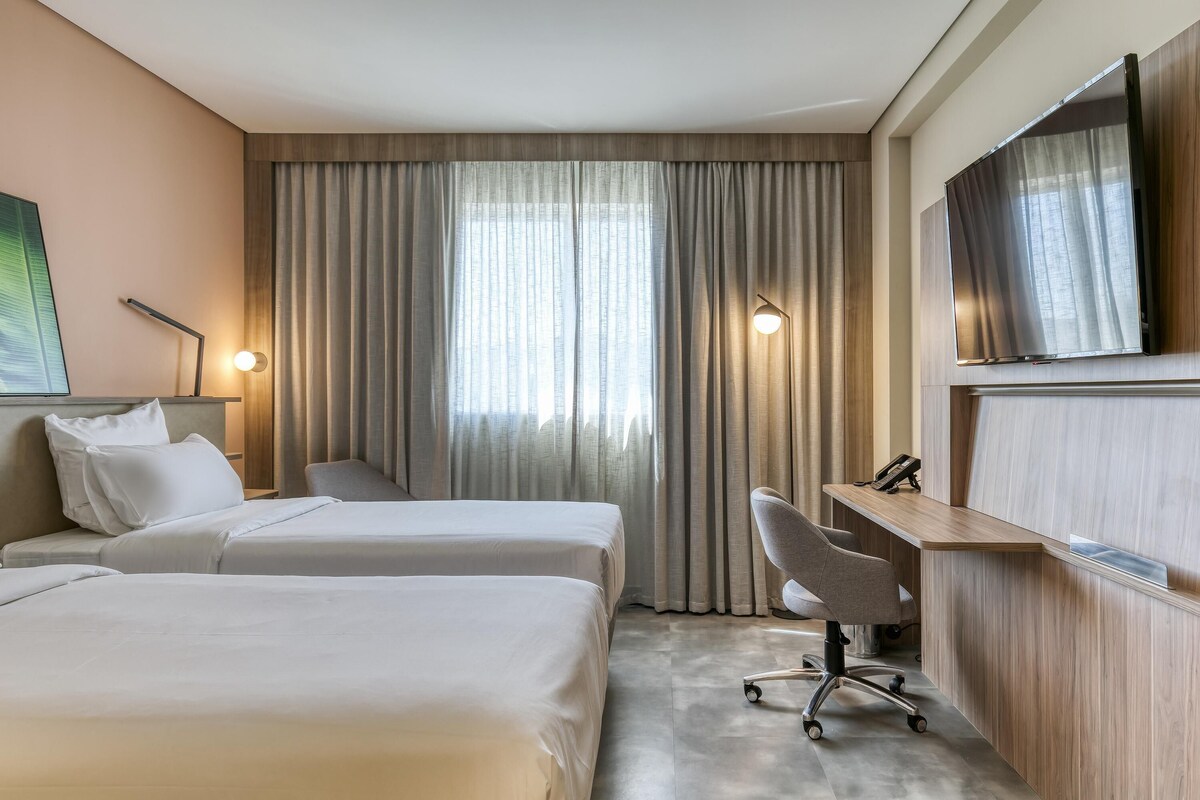 Une chambre d’hôtel avec deux lits, un bureau, une chaise, une télévision et une grande fenêtre avec des rideaux.
