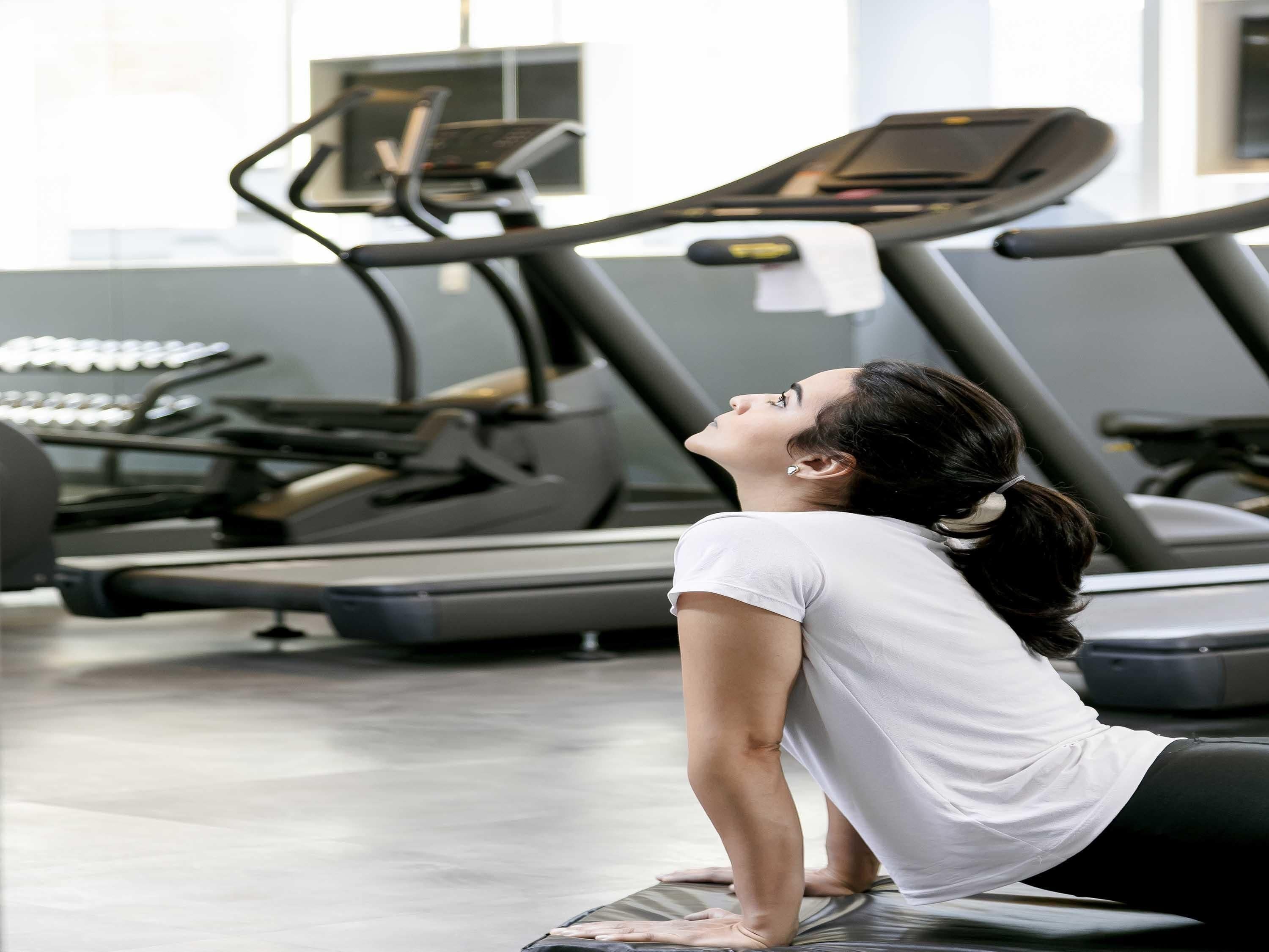 Una persona realizando una postura de yoga sobre una colchoneta en un gimnasio, con cintas de correr al fondo.
