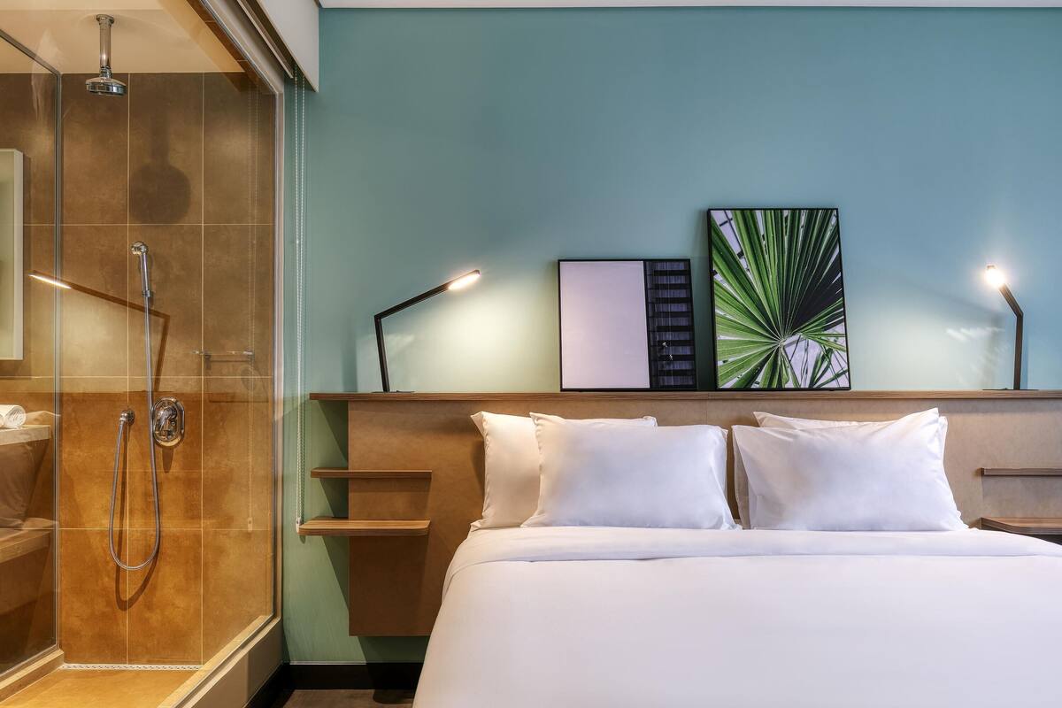 Habitación de hotel con ducha, cama con ropa blanca, cabecero con estantes y dos lámparas fijadas a la pared.