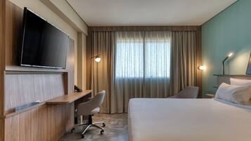 Quarto de hotel com cama, escrivaninha, cadeira, televisão e cortinas.
