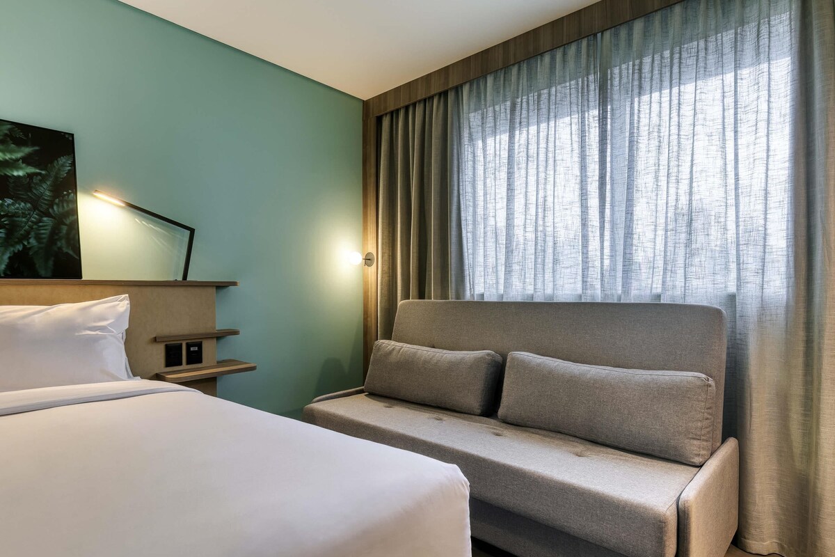 Une chambre d’hôtel avec un lit, un canapé, une étagère fixée au mur et une fenêtre avec des rideaux.