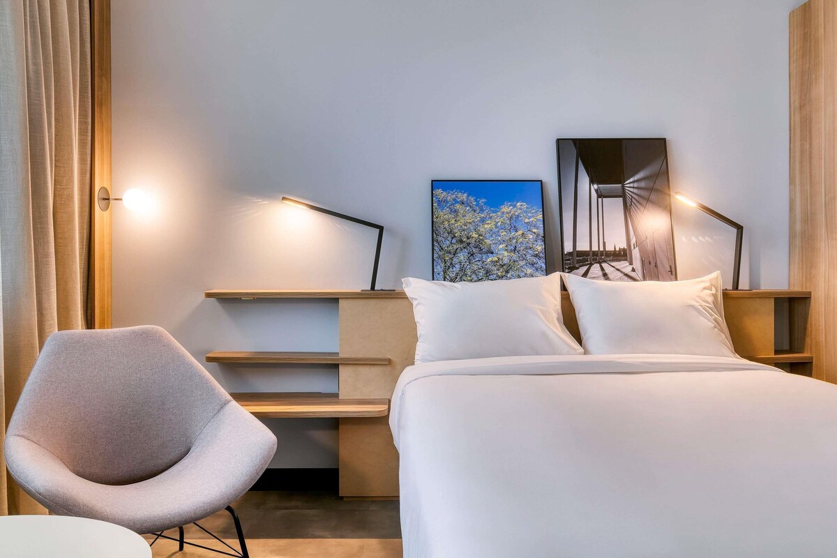 Une chambre d’hôtel moderne, équipée d’un lit, d’une chaise, d’un bureau et d’un tableau au mur.