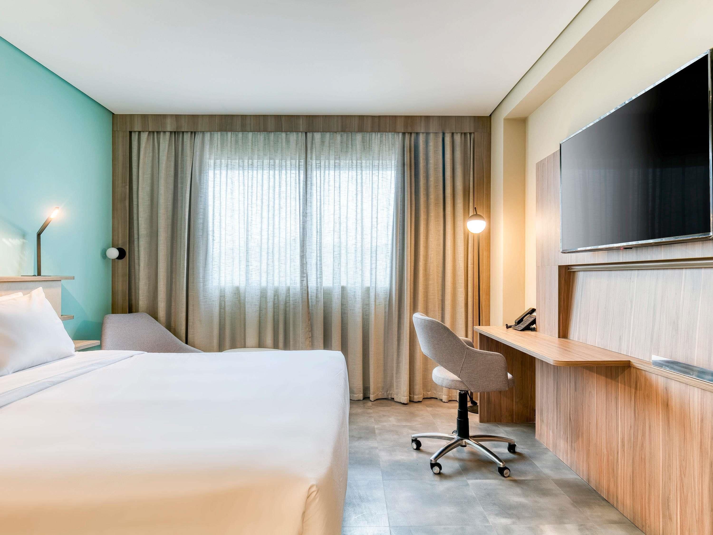 A hotel room with a bed, a desk, a chair, a TV, and a window with curtains.