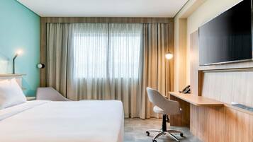A hotel room with a bed, a desk, a chair, a TV, and a window with curtains.