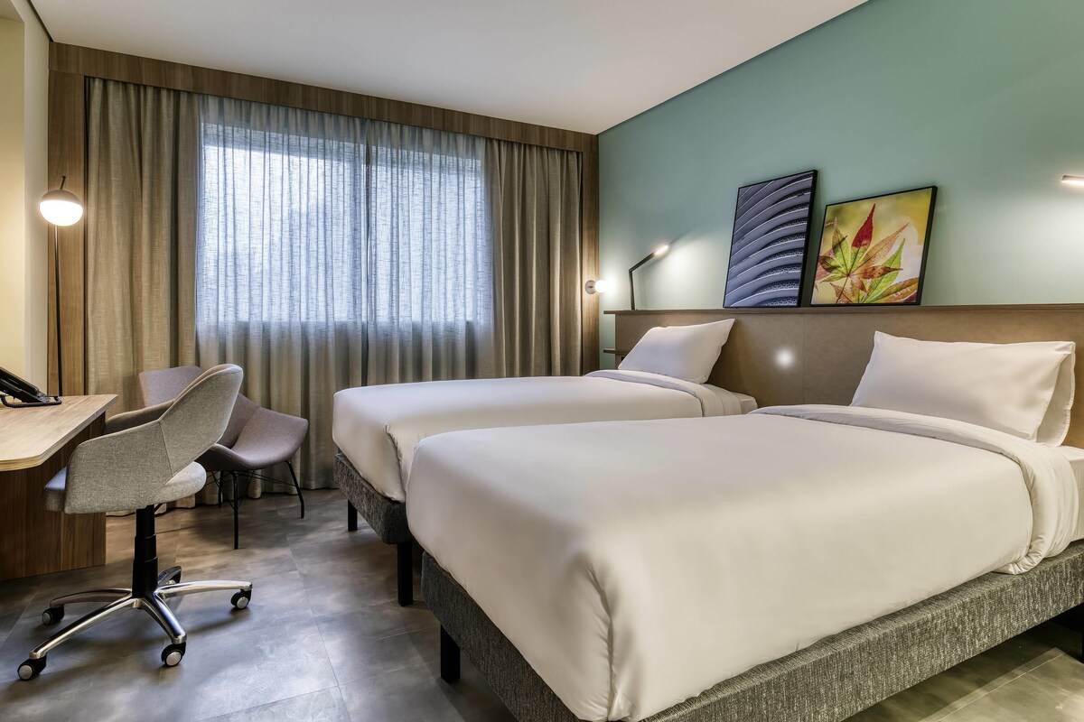 A hotel room with two beds, a desk with a chair, a lamp, and a window with curtains.