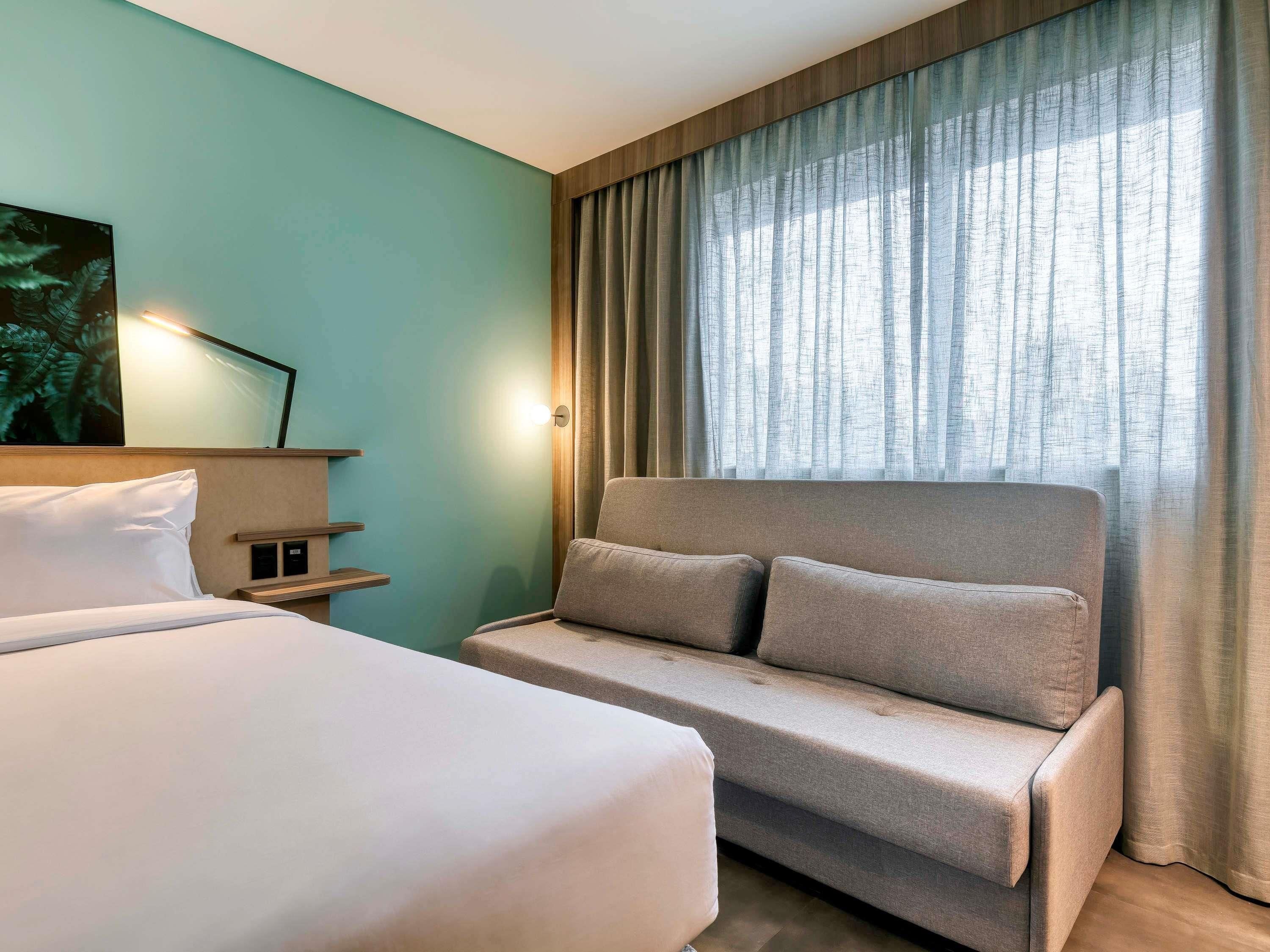 A hotel room with a bed, a sofa, a wall-mounted shelf, and a window with curtains.