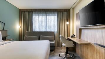 Una habitación de hotel con una cama grande, un escritorio con una silla, un televisor de pantalla plana y un ventanal con cortinas.