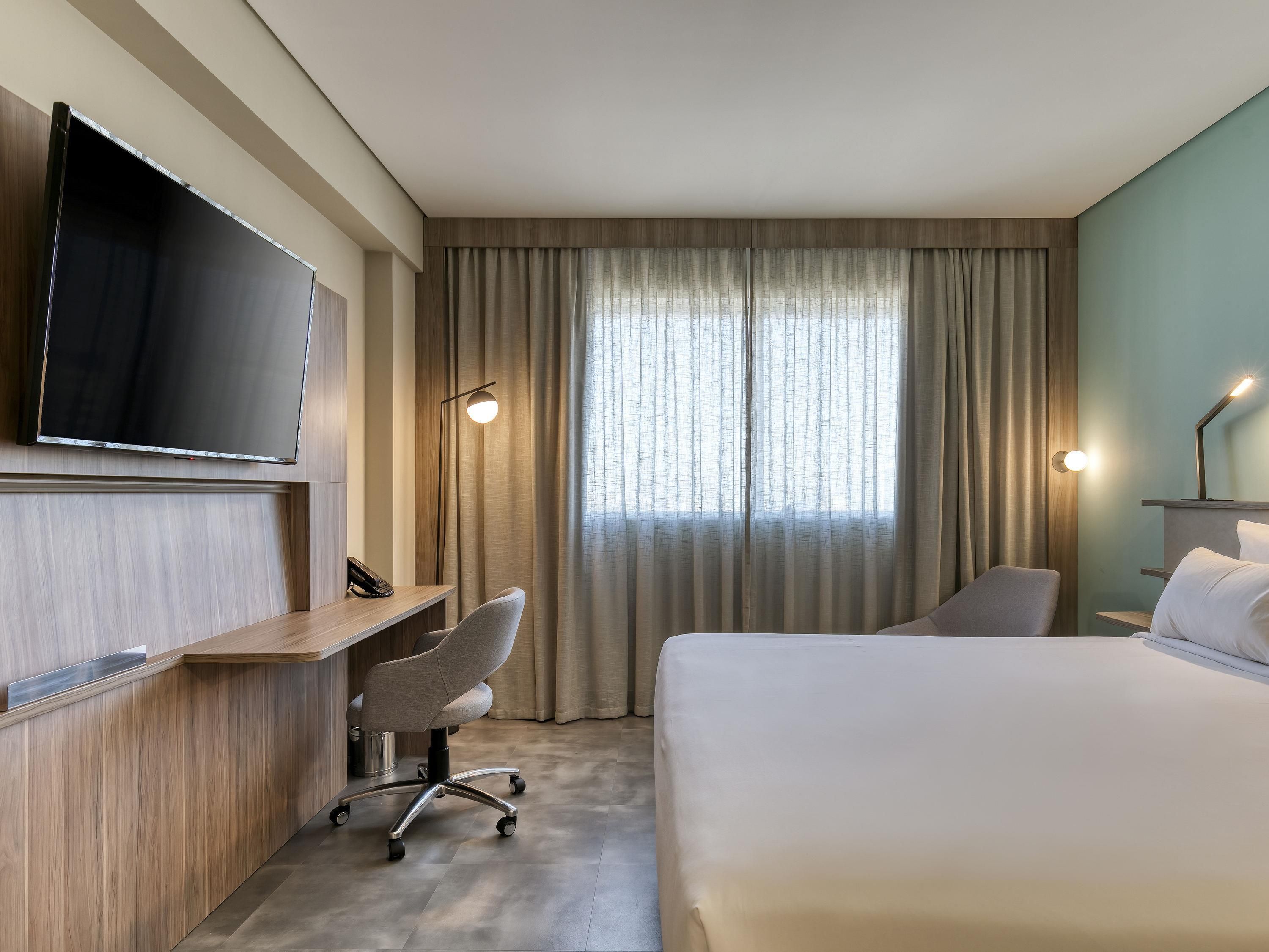 Quarto de hotel com cama, escrivaninha, cadeira, televisão e cortinas.