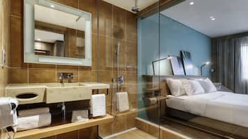 Un baño de hotel moderno con ducha de vidrio, un espejo grande, un lavamanos con toallero y una cama con ropa blanca.