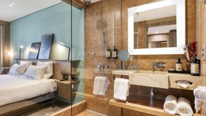 Een moderne hotelbadkamer met een douchecabine van glas, een grote spiegel, een wastafel met toiletartikelen en een bed met witte lakens.