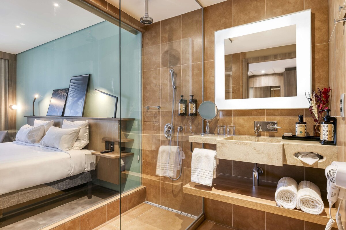 Une salle de bain d’hôtel moderne avec une douche en verre, un grand miroir, un meuble-lavabo avec des articles de toilette et un lit avec du linge de maison blanc.