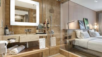 Un baño de hotel moderno con ducha de vidrio, un espejo grande, tocador con artículos de aseo y cama con ropa blanca.