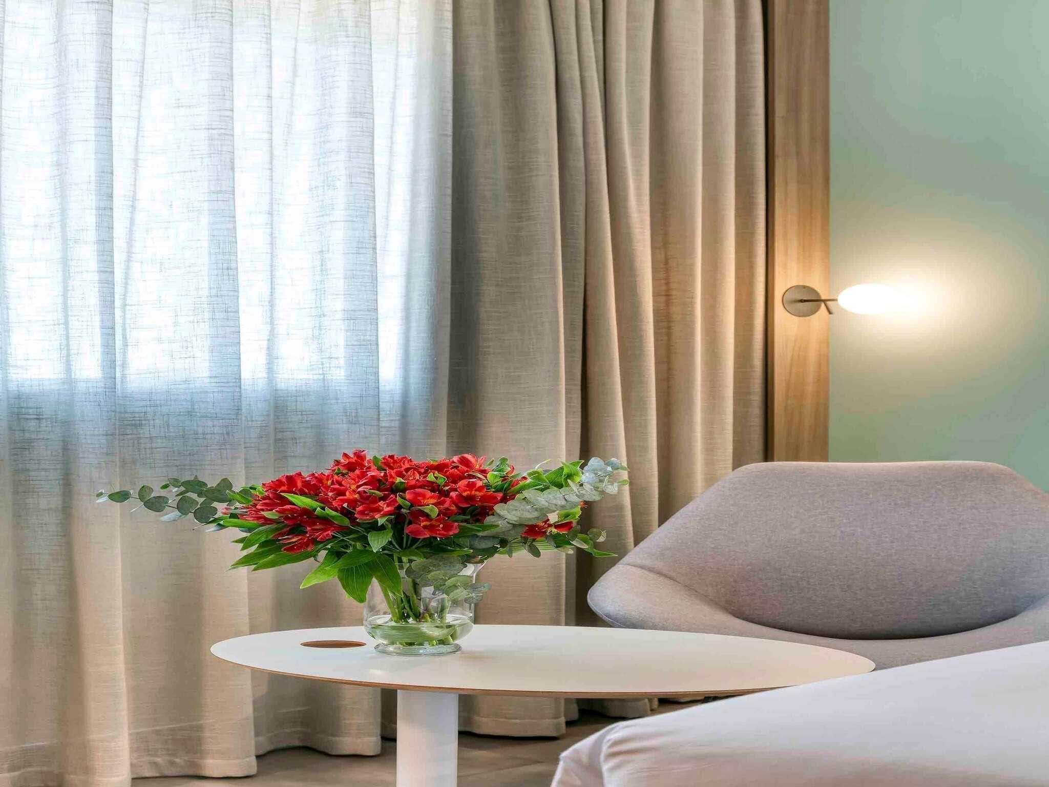 A room with a round table, a chair, a vase with flowers, and curtains.