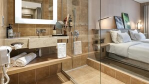 Habitación de hotel moderna con cabina de ducha de vidrio, un espejo grande, mesita de noche con artículos de aseo y cama con ropa de cama blanca.
