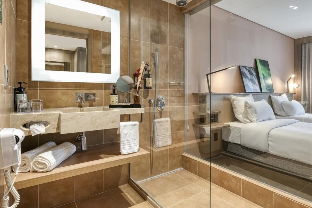 Une chambre d’hôtel moderne dotée d’une douche en verre, d’un grand miroir, d’une table de chevet avec des articles de toilette et d’un lit avec une literie blanche.