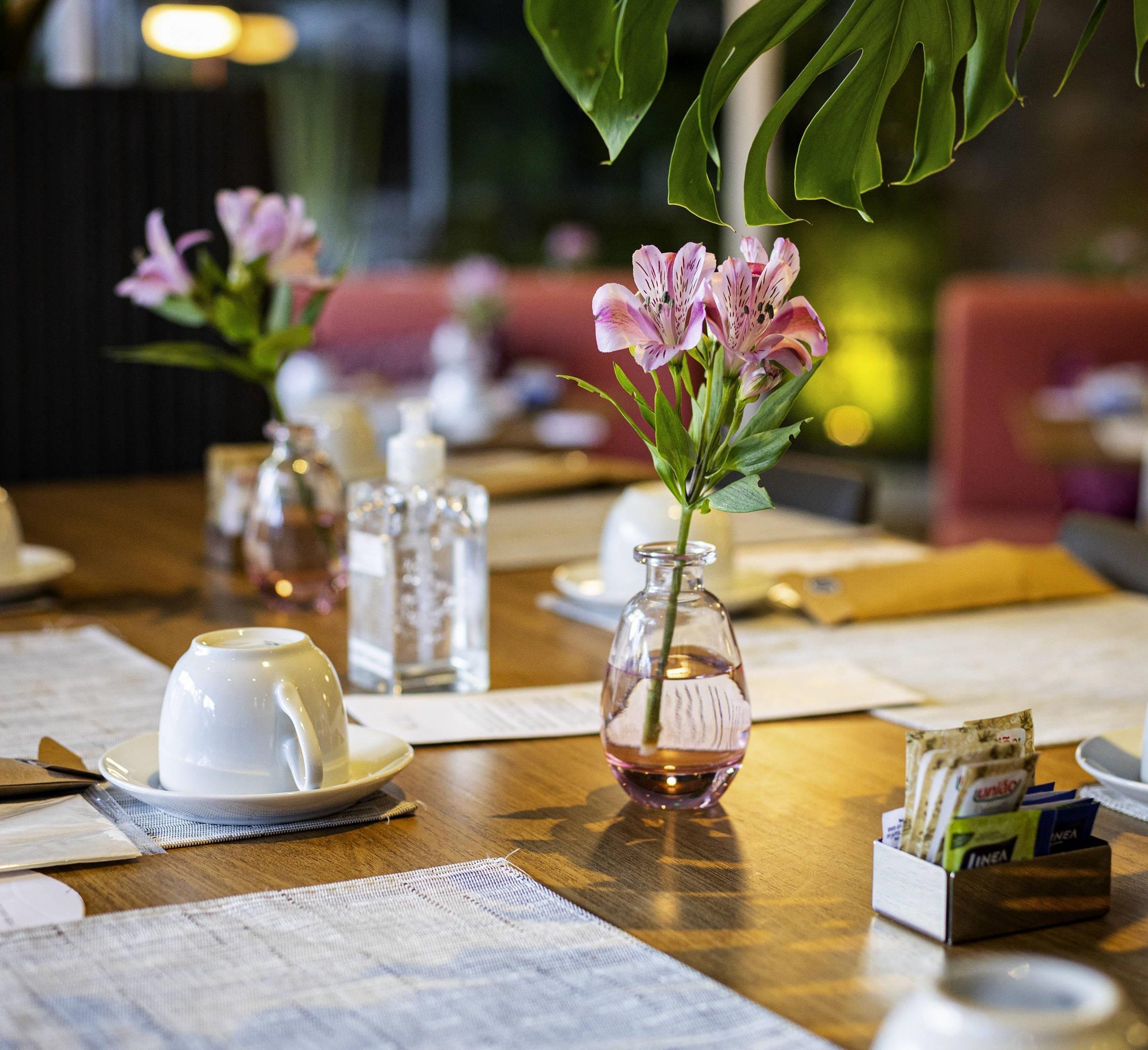 A dining table with a white teapot, a glass vase with pink flowers, and a water bottle.