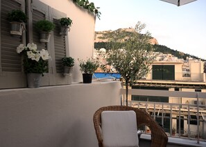 Varanda com plantas em vasos, uma cadeira de vime e vista para uma colina com edifícios.